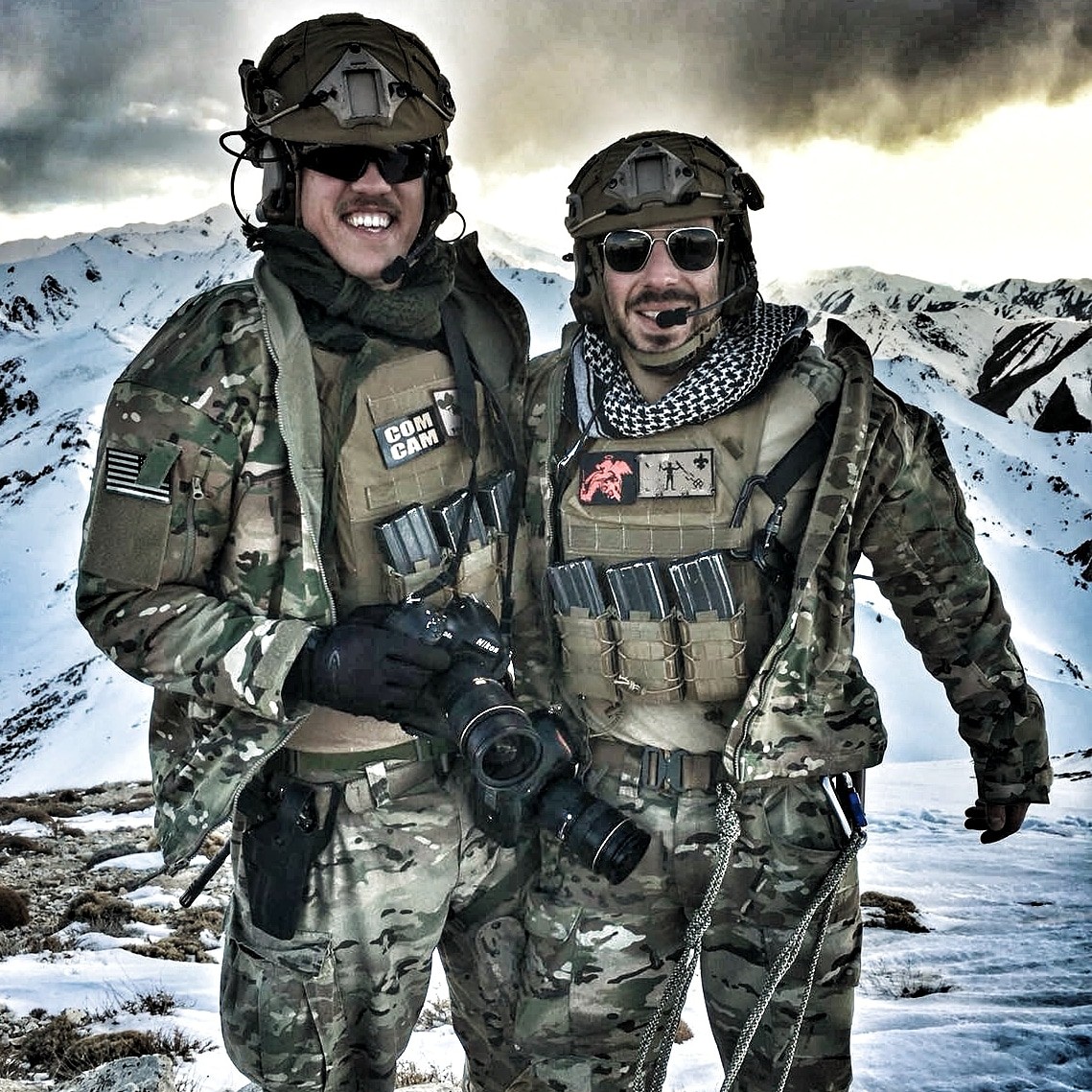 Gregory Brook, wearing military gear, poses for picture with fellow service member in front of a mountain range.