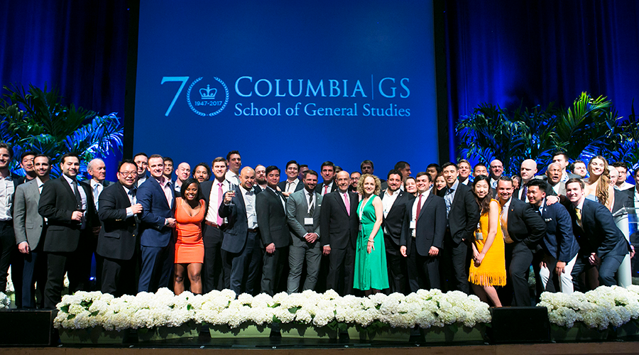 Veteran attendees to the Annual GS Alumni Dinner gather on stage with Dean Peter J. Awn, with the 70th anniversary GS logo projected behind them
