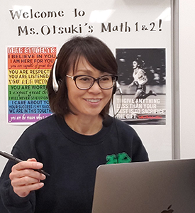 Mayumi Otsuki sitting at laptop, teaching, with inspiring words to students in the backround