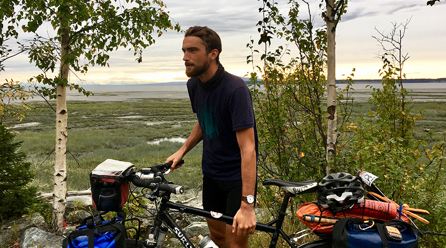 Man leaning on mountain bike loaded with gear, standing in front of two birch trees and marshland