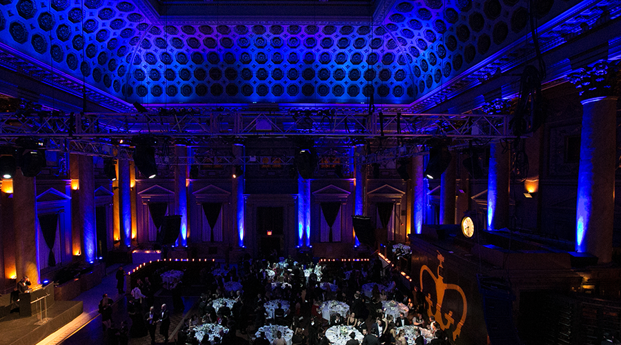 Picture of a large, blue-lit room with high, ornate ceilings looking down at round tables with people sitting