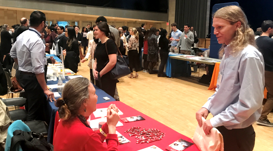 A student speaks with an admissions representative at their table in the foreground, with dozens of students in the background speaking to other representatives